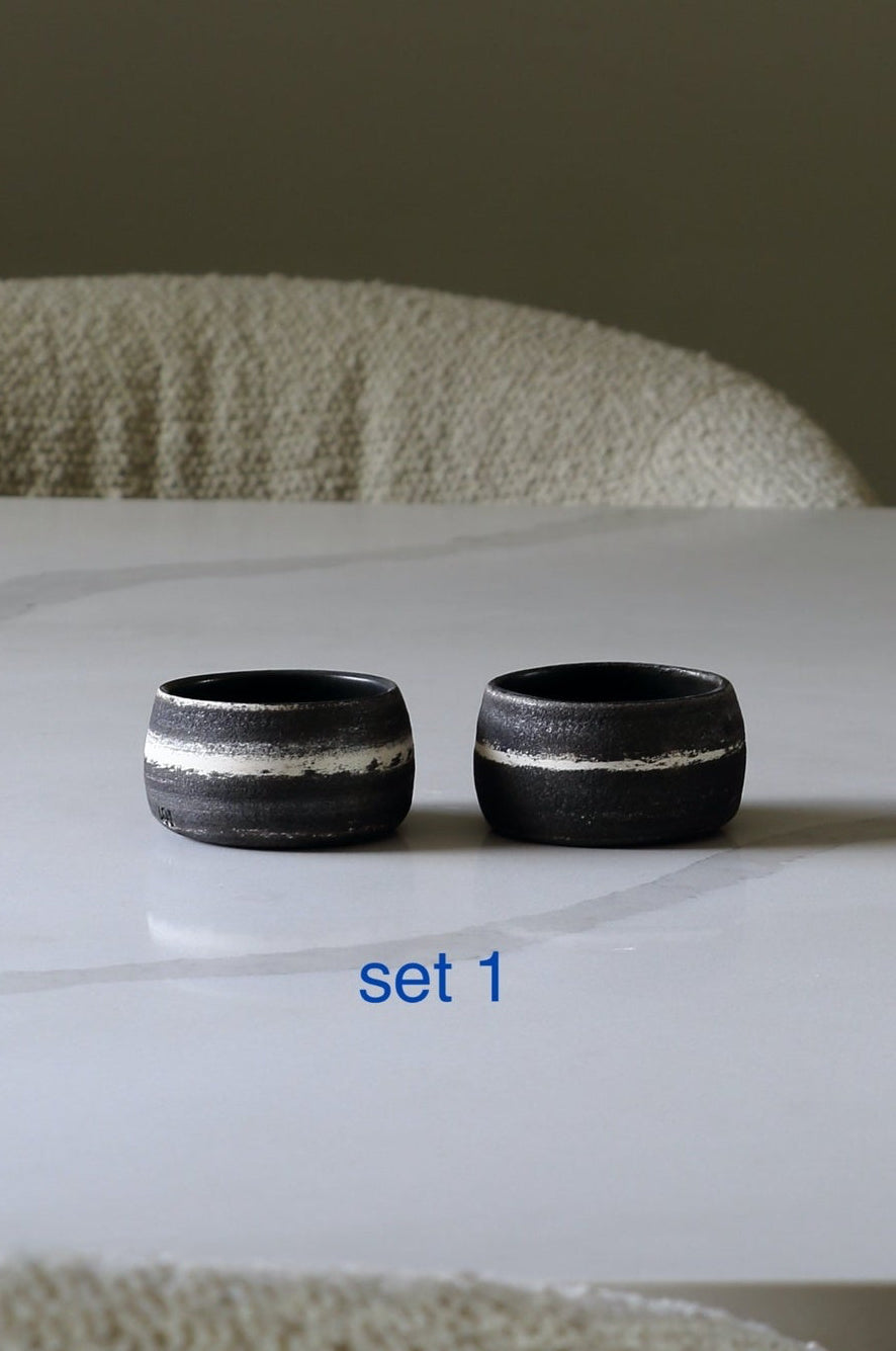 Two small dark ceramic bowls with a white horizontal band, placed on a white table with textured beige chairs in the background. Blue text below reads 'set 1'.