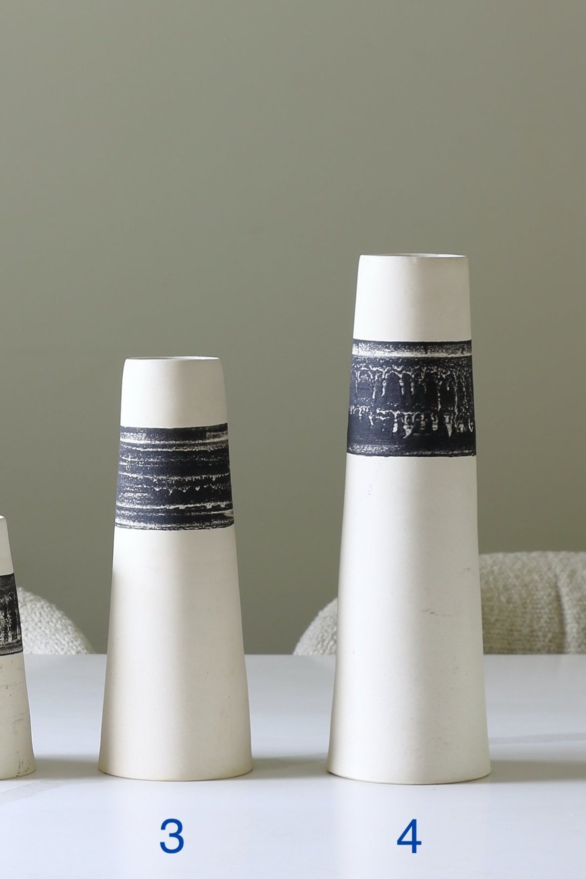 Two white ceramic vase-like containers with dark navy patterned bands, shown on a tabletop against a muted green-gray background. Blue numbers 3 and 4 label the two vases.
