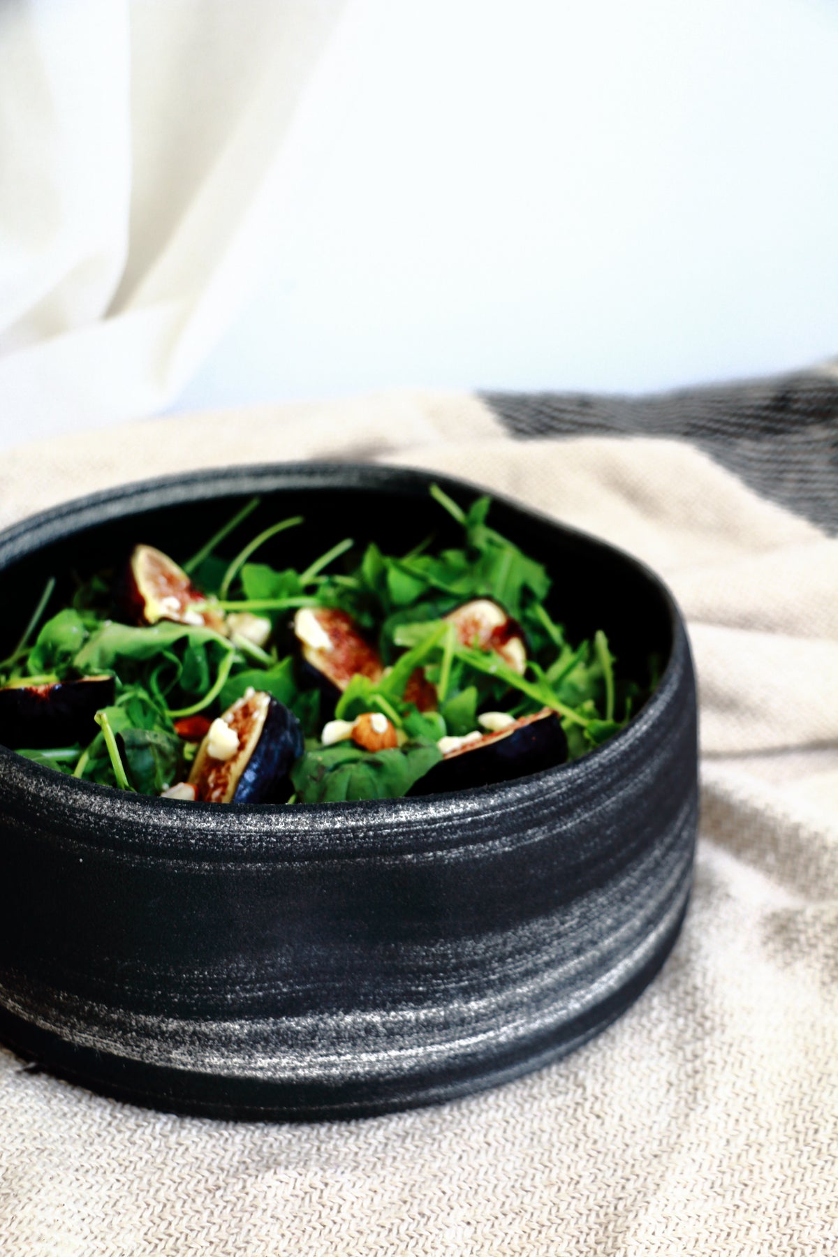 A black textured ceramic bowl filled with a green salad containing figs and nuts, set on a beige linen cloth.