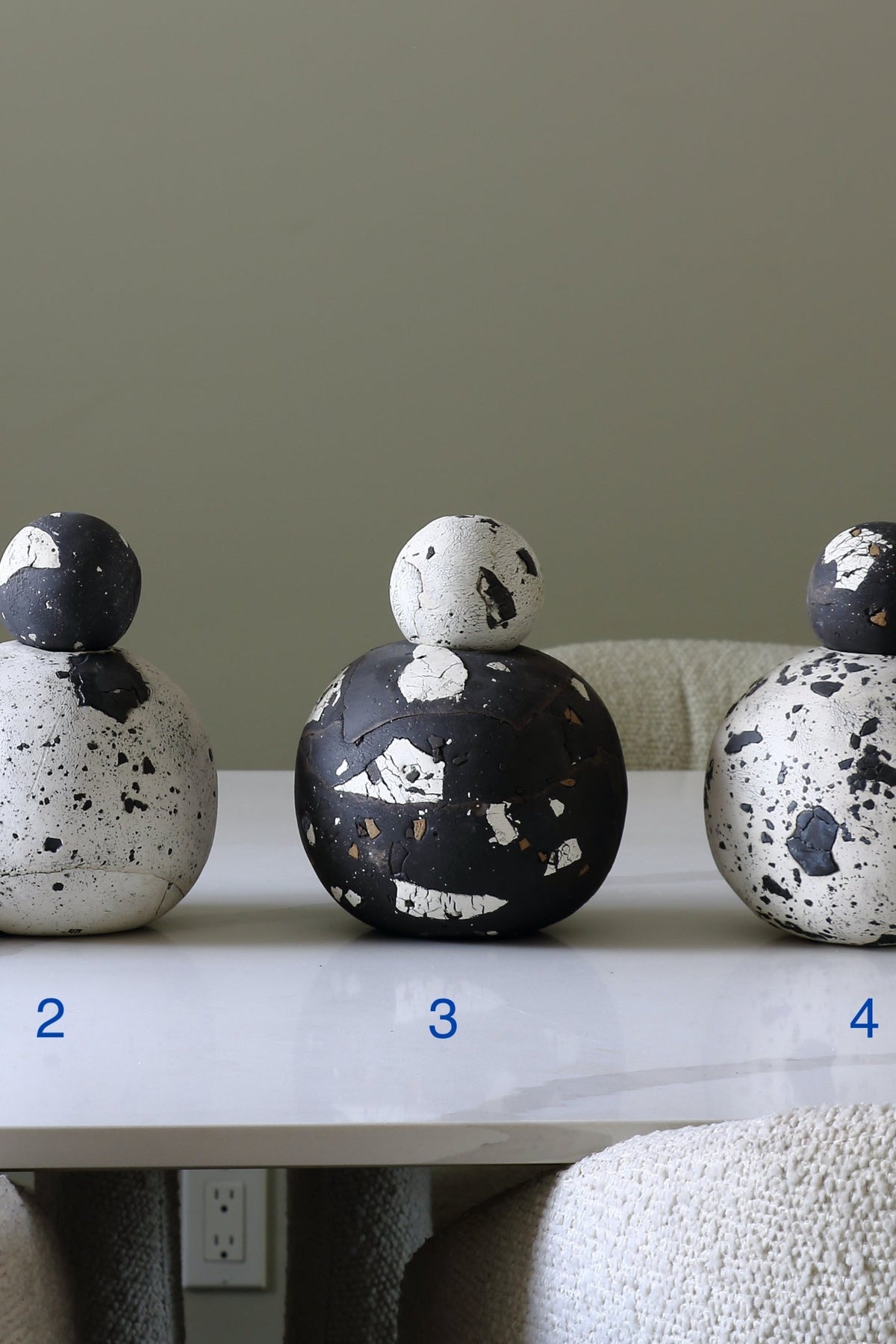 Three decorative ceramic vases with rounded, sculptural forms and black-and-white terrazzo-style speckling, displayed on a white table against a muted green wall. Blue numbers 2, 3, and 4 appear below each vase.