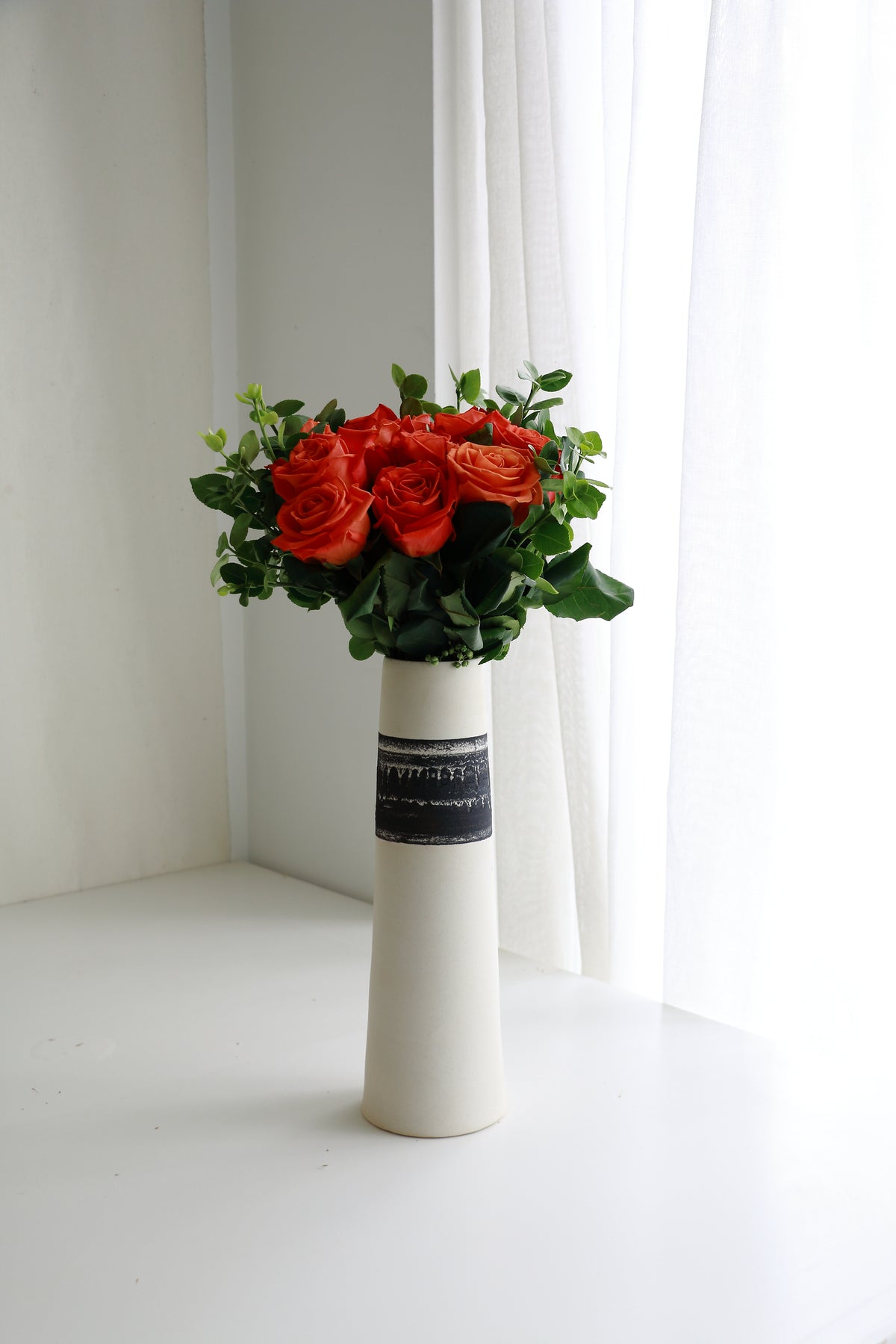 A bouquet of red roses with green leaves arranged in a tall white ceramic vase with a black patterned band, set on a white surface near a window with sheer curtains.
