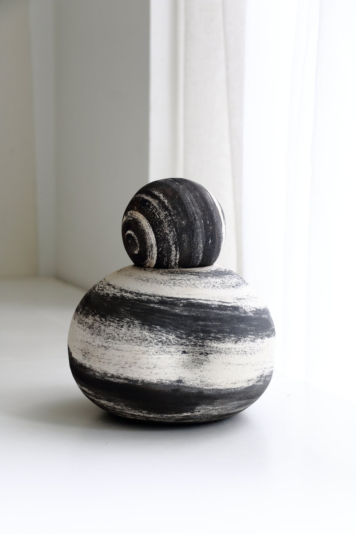 A minimalist ceramic sculpture of a snail, featuring a textured black and white striped round base with a matching striped dome shell on top, set against a bright, softly lit white background.