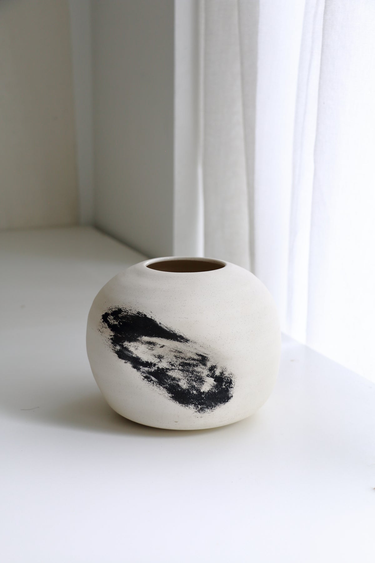 A small round ceramic vase with a matte off-white finish and a bold black abstract brushstroke on the front, set on a white windowsill with soft natural light from a nearby window.