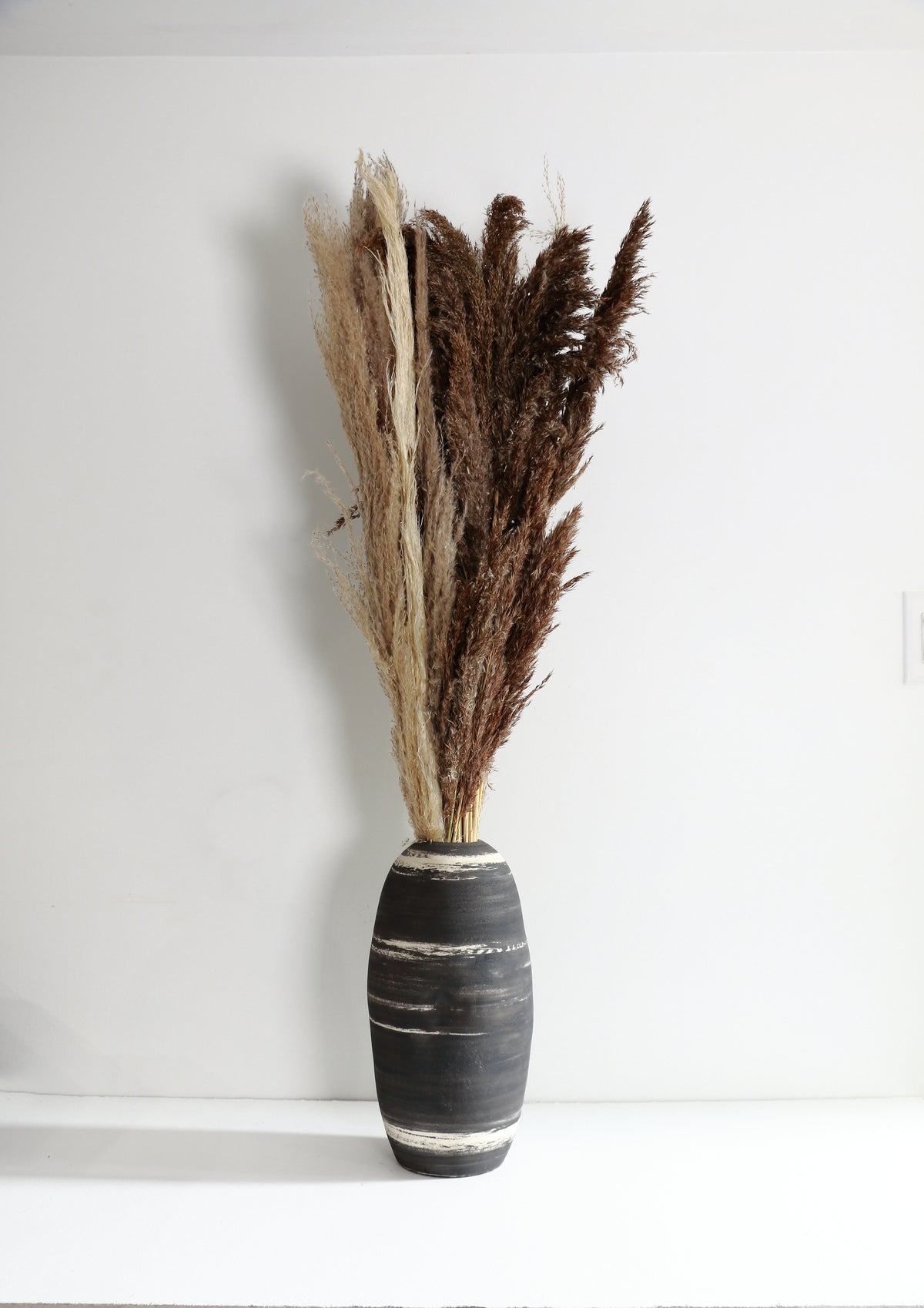 A dried pampas grass arrangement in a dark gray ceramic vase with white painted stripes, set against a plain white wall and surface.