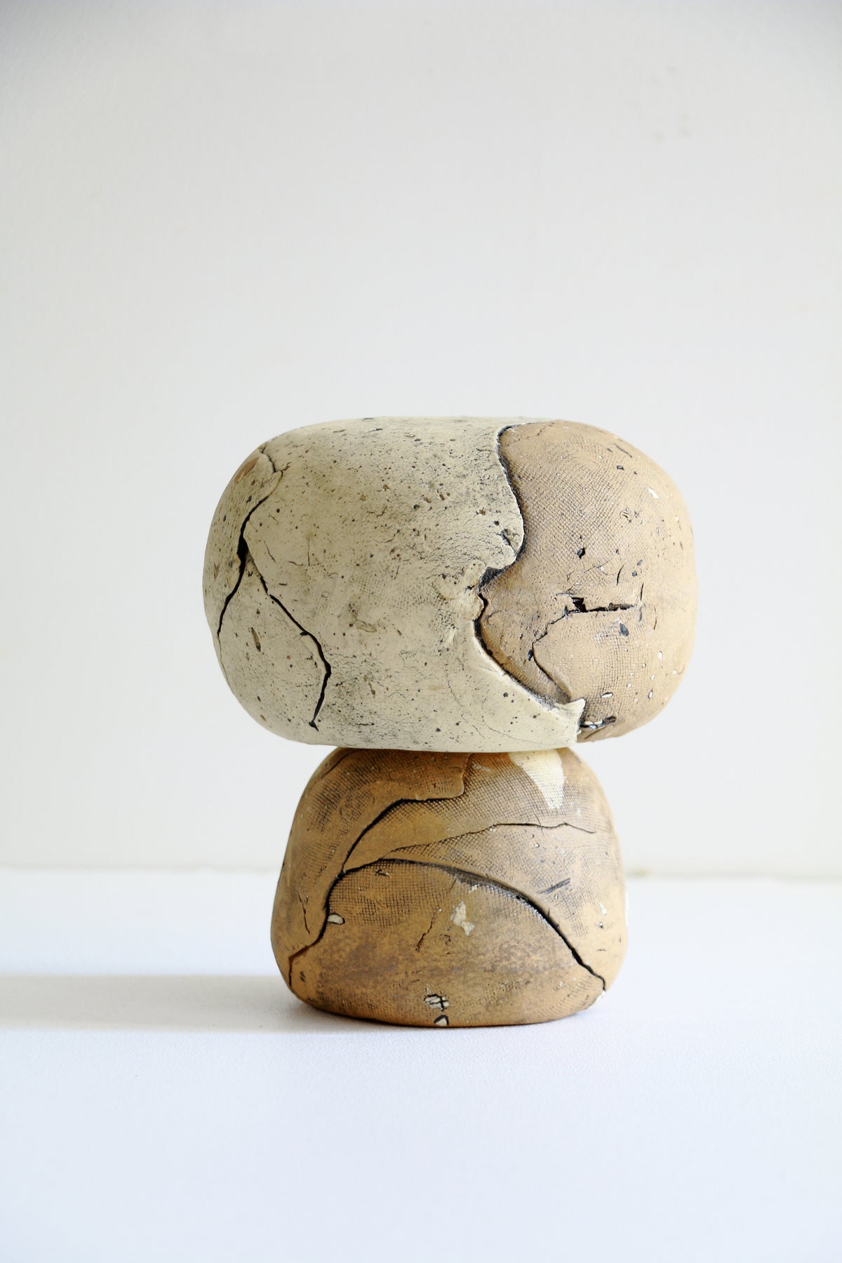 A small sculptural object made of two stacked, cracked stone-like pieces, one light beige and one warm brown, photographed against a plain white studio background.