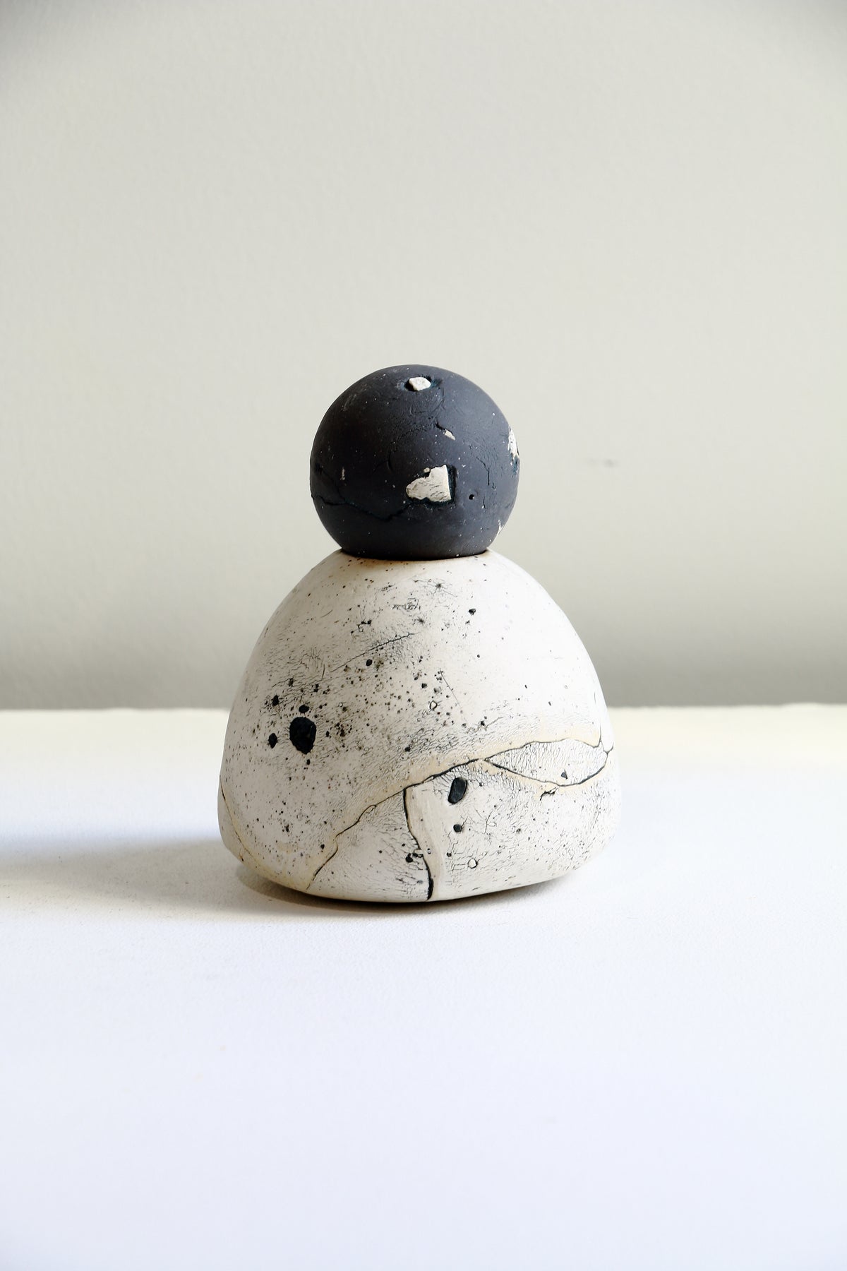 A small handmade ceramic sculpture with a matte black spherical top and a speckled off-white rounded base, photographed on a plain light background.