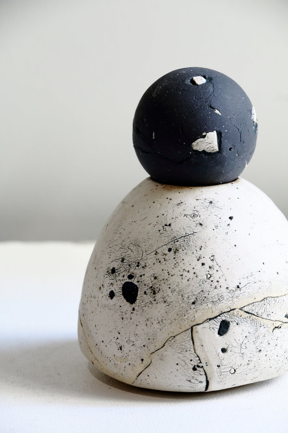 A sculptural ceramic object featuring a matte black spherical top resting on a textured, speckled off-white dome base, photographed against a neutral light gray background.