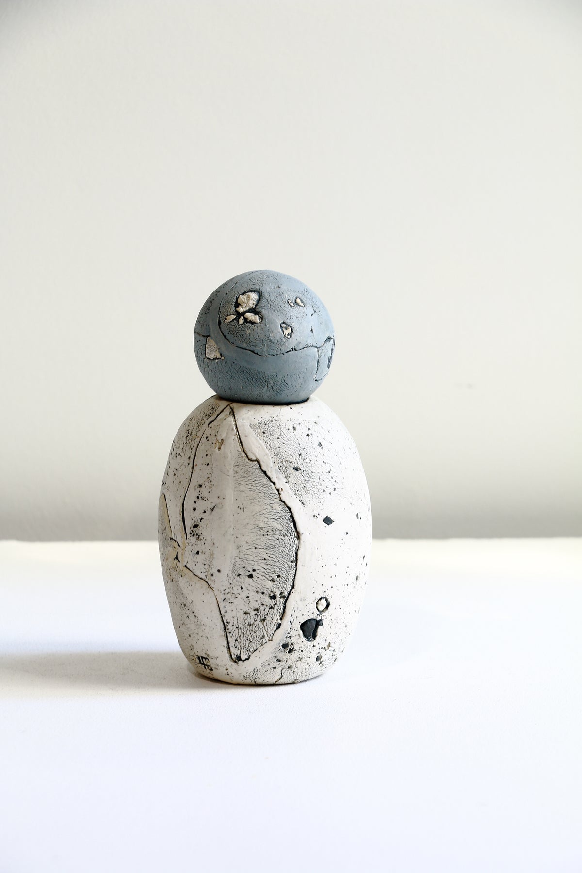A small handmade ceramic figurine resembling a penguin, featuring a speckled off-white body and a muted blue-gray spherical head, set against a plain light background.