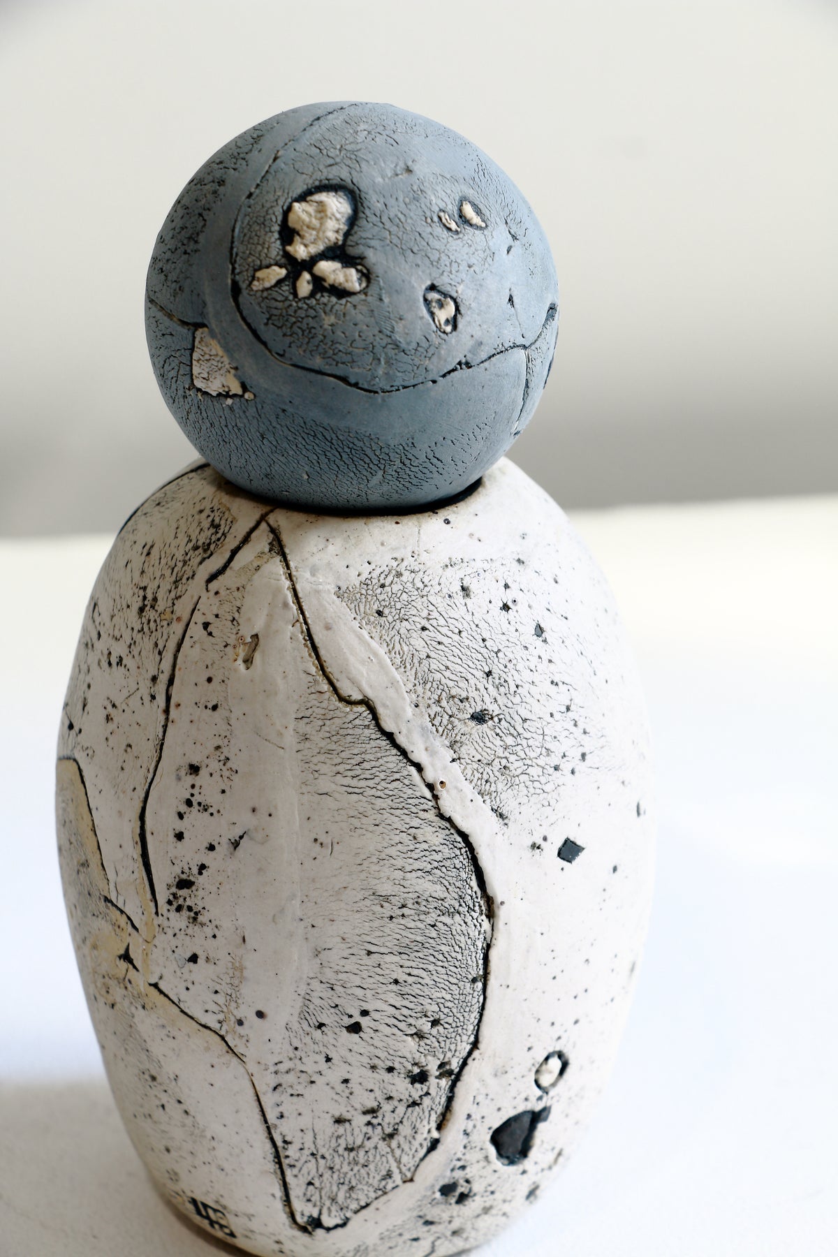 A handmade ceramic sculpture of a stylized figure, featuring a textured, speckled off-white body and a smooth, matte slate-blue spherical head, set against a plain light background.