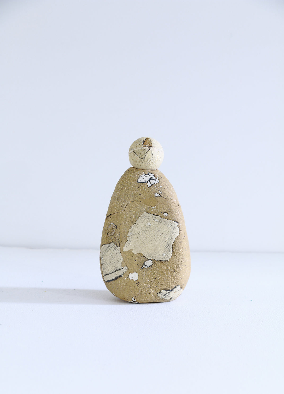 A small handmade ceramic figurine with a textured, organic shape in beige and cream, featuring peeling white and gray clay layers, standing on a white surface against a white background.