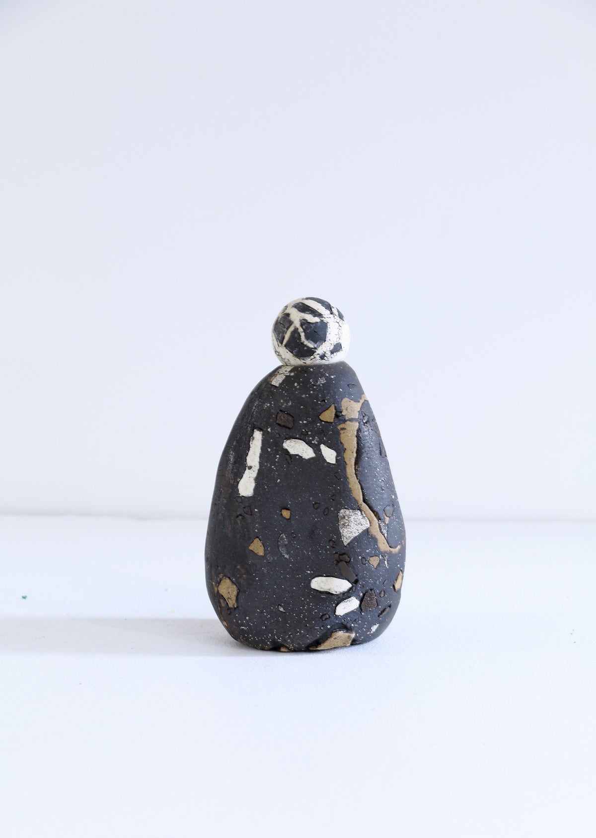 A small handmade ceramic figurine shaped like a stylized person, featuring a dark charcoal body with white and gold speckled details, standing on a white surface against a white background.