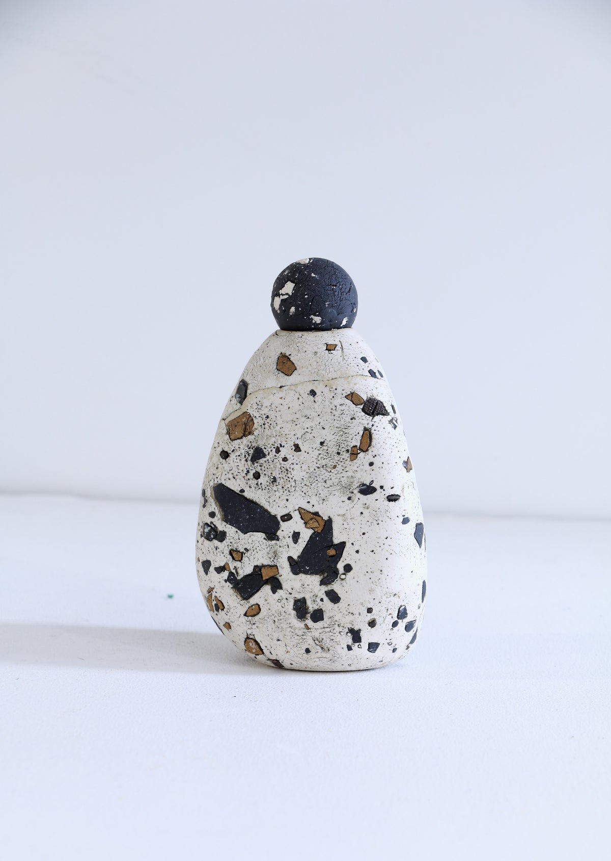A small handmade ceramic bottle with a rounded teardrop shape, featuring a speckled off-white body with black and brown flecks, topped with a dark charcoal black spherical lid, set against a plain white background.