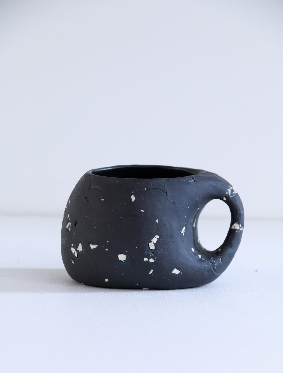 A matte black ceramic mug with a distinctive loop handle and white terrazzo-like flecks, photographed on a clean white surface against a light gray background.