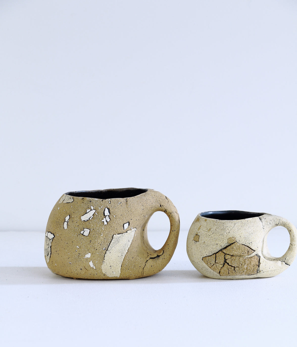 Two handmade ceramic mugs with a textured, earthy finish and a crackled glaze, displayed on a clean white surface against a light gray background.