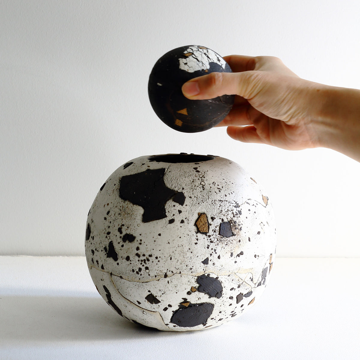 A hand holds a small black and white textured ceramic orb above a larger matching ceramic vase with a similar black and white marbled pattern, set against a plain white background.
