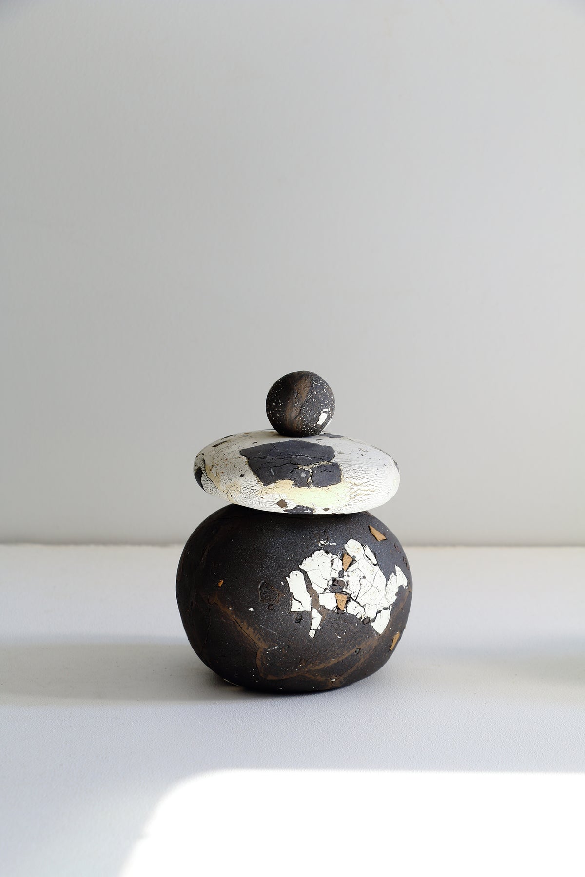 A small handmade ceramic lidded container with a rounded dark body and lid, featuring distressed white and black paint details, set against a plain light gray background.