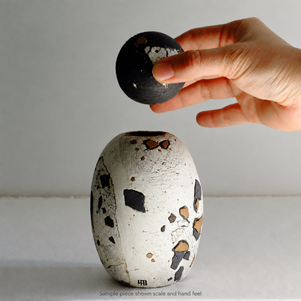 A hand holds a small black round object above a textured white ceramic vase with black and brown speckles on a plain light background.