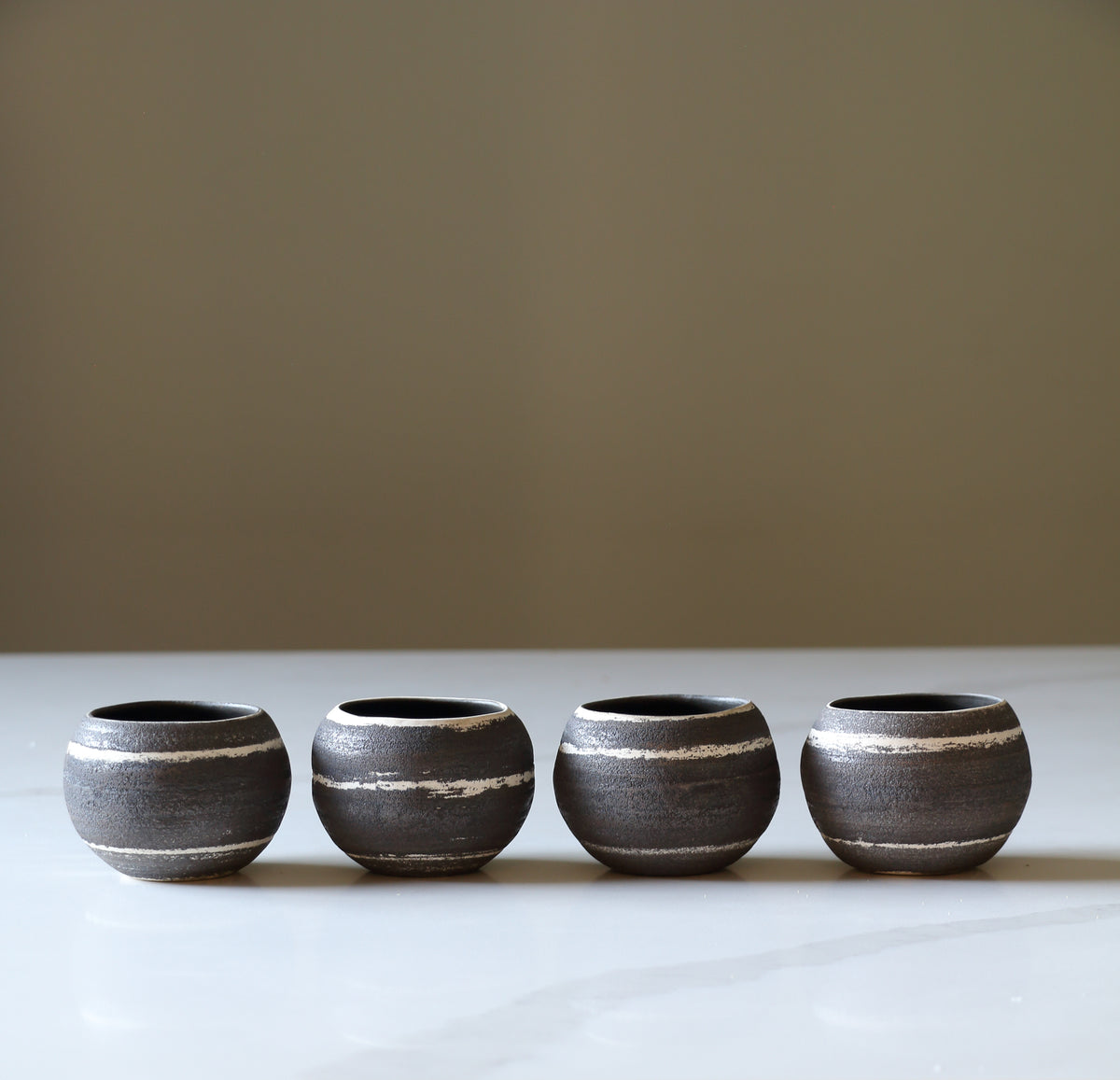 Four small round ceramic cups with a dark charcoal and beige horizontal striped pattern, lined up on a light surface against a solid neutral background.