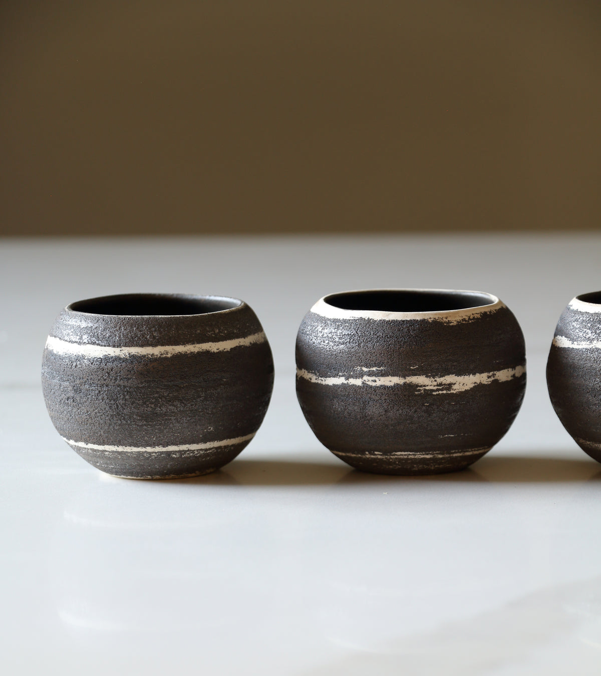 Three small round ceramic cups with a dark charcoal-brown glaze and horizontal distressed white stripes, arranged on a light surface against a warm neutral background.