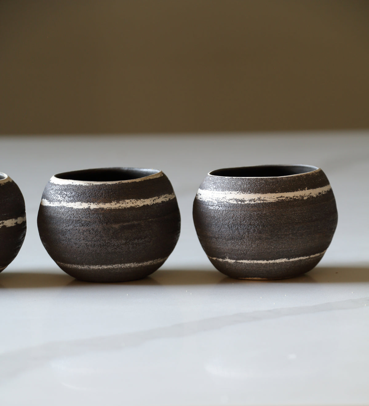 Three small round ceramic cups with a dark textured glaze and horizontal light beige stripes, arranged on a light surface against a neutral background.