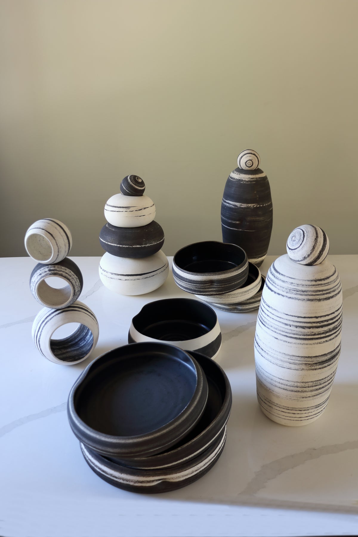 A collection of handmade ceramic tableware and decorative vases in black, white, and gray striped glazes, arranged on a light marble surface against a neutral beige wall.