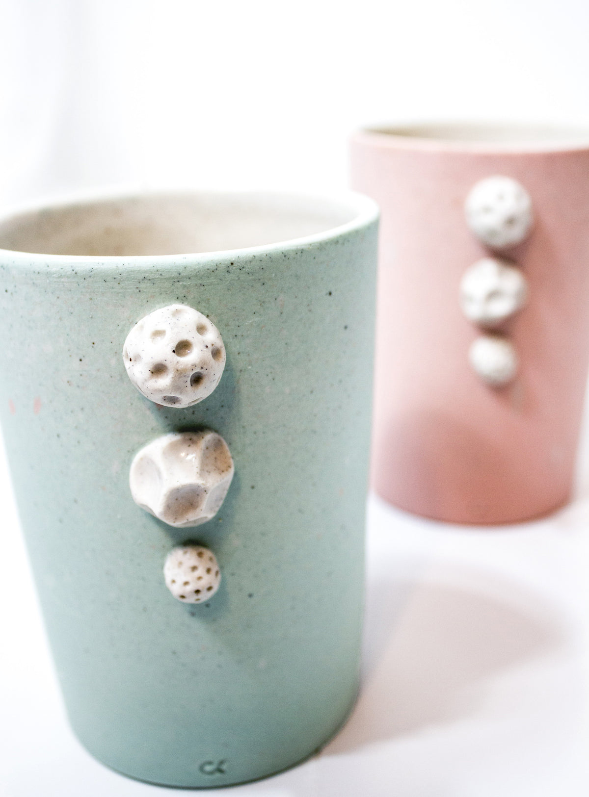 Two handmade ceramic mugs with organic, raised stone-like accents, one in sage green and one in dusty pink, set against a bright white background.
