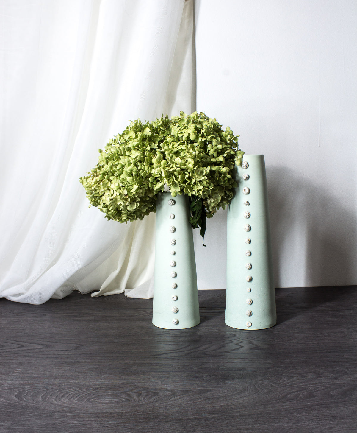 Two pale mint green ceramic vases with vertical rows of metallic studs, one holding a large green hydrangea bouquet and the other empty, set on a dark wood floor against a white curtain and wall.