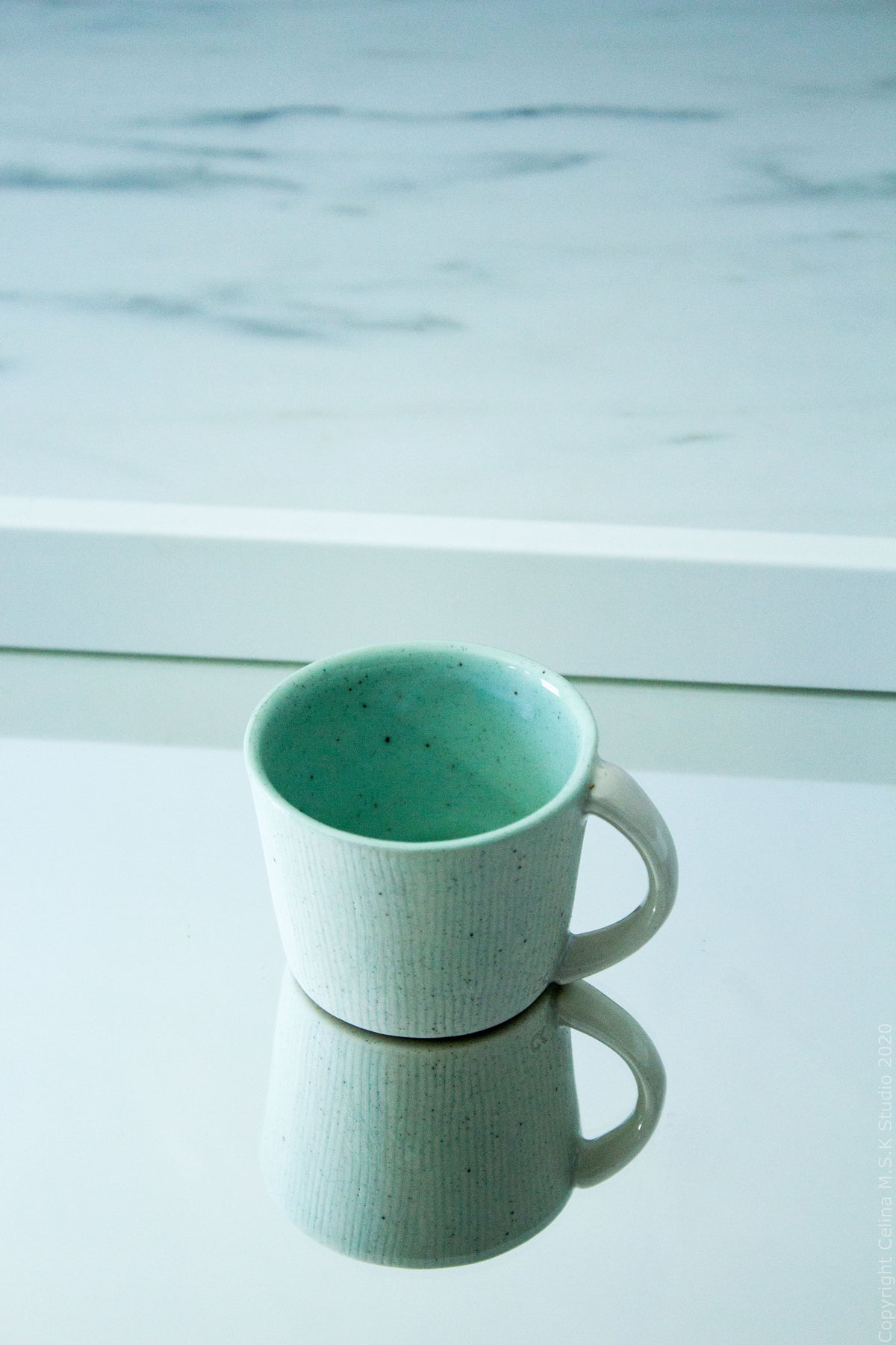 A small ceramic mug with a speckled mint green interior and a light gray-green exterior, shown on a reflective white surface with a blurred light blue background.