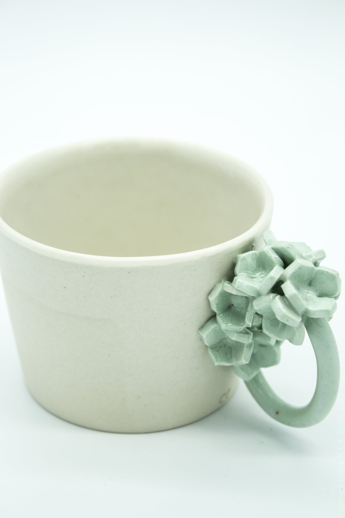 A light beige ceramic mug with a sculptural, textured pale green handle shaped like a cluster of flowers, set against a plain white background.