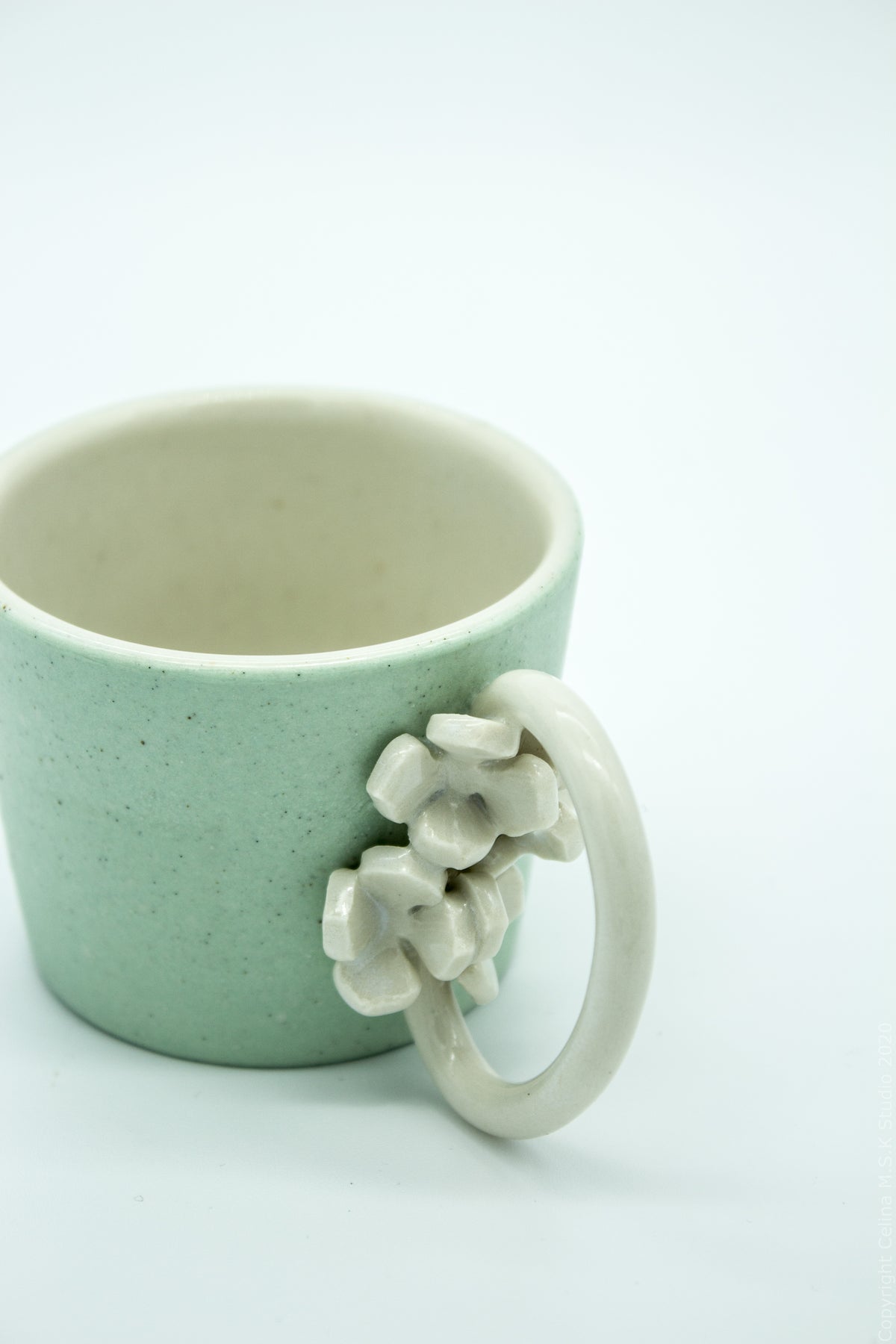 A speckled light green ceramic mug with a cream-colored handle decorated with sculpted flowers, set against a plain white background.