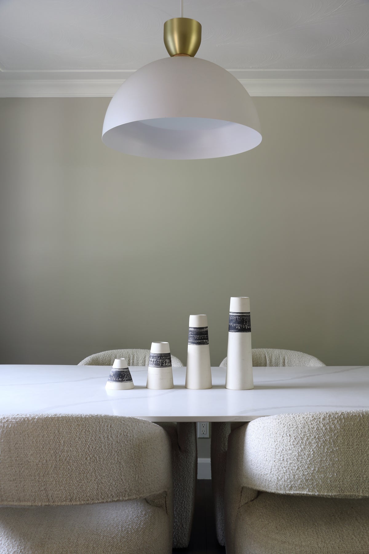 A modern white dome pendant light with a brushed gold ceiling mount hanging over a dining table with textured cream boucle chairs and small decorative ceramic vases.