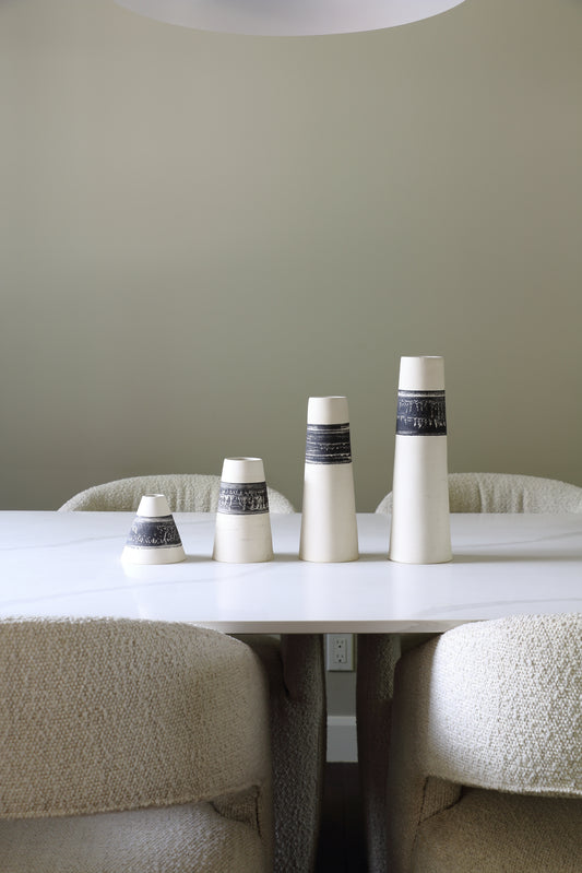 Four white ceramic vases with dark gray patterned bands, arranged in ascending size on a white dining table between textured cream boucle chairs.