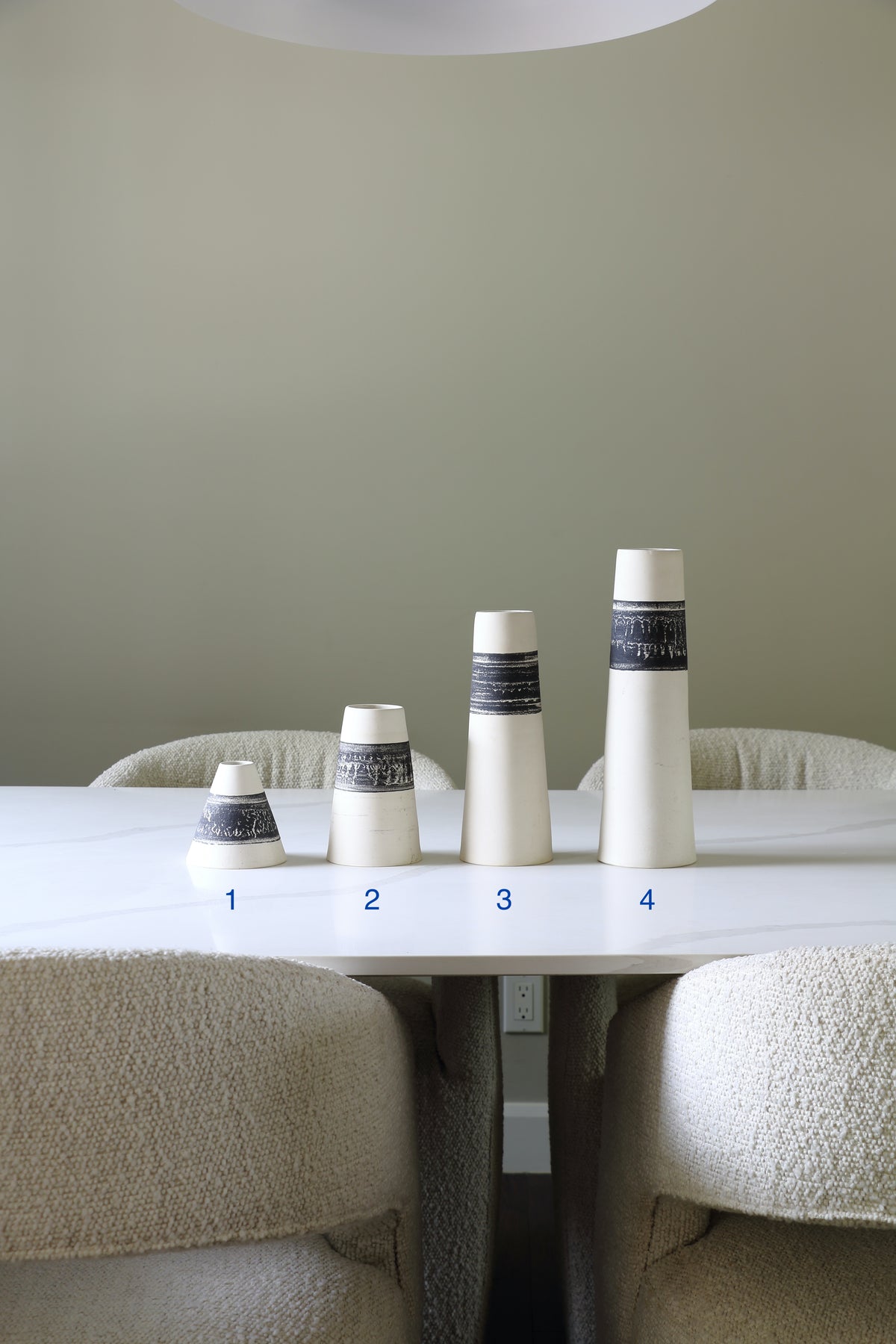Four tapered ceramic cups with a white body and a dark navy patterned band, arranged by size on a white dining table. The numbers 1 through 4 are printed in blue near the base of each cup.