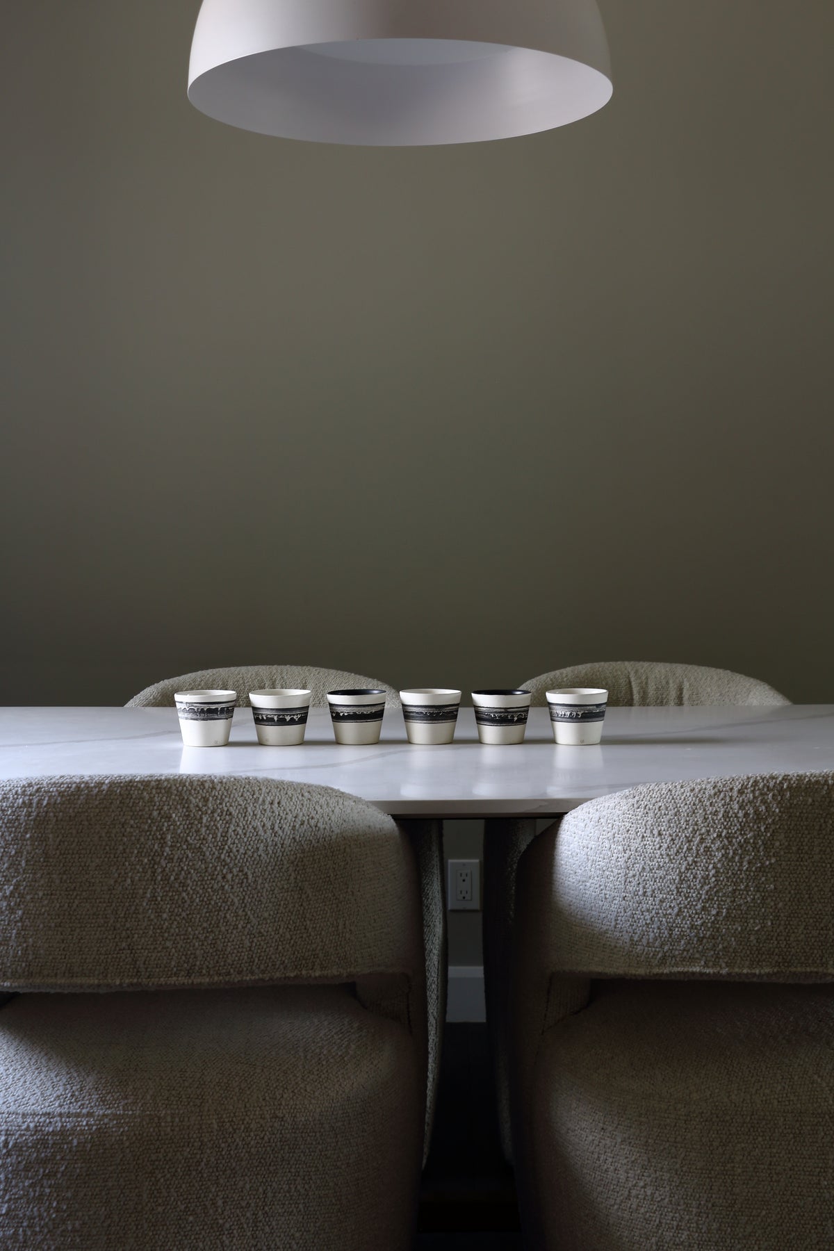 A dining table with six small metallic cups lined up on top, surrounded by upholstered chairs in a minimalist interior with a large white pendant light overhead.