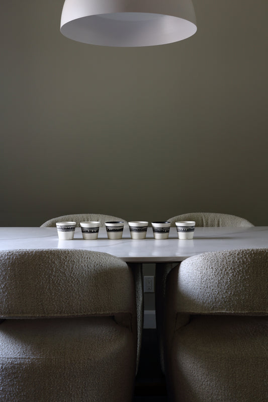 A dining table with six small metallic cups lined up on top, surrounded by upholstered chairs in a minimalist interior with a large white pendant light overhead.