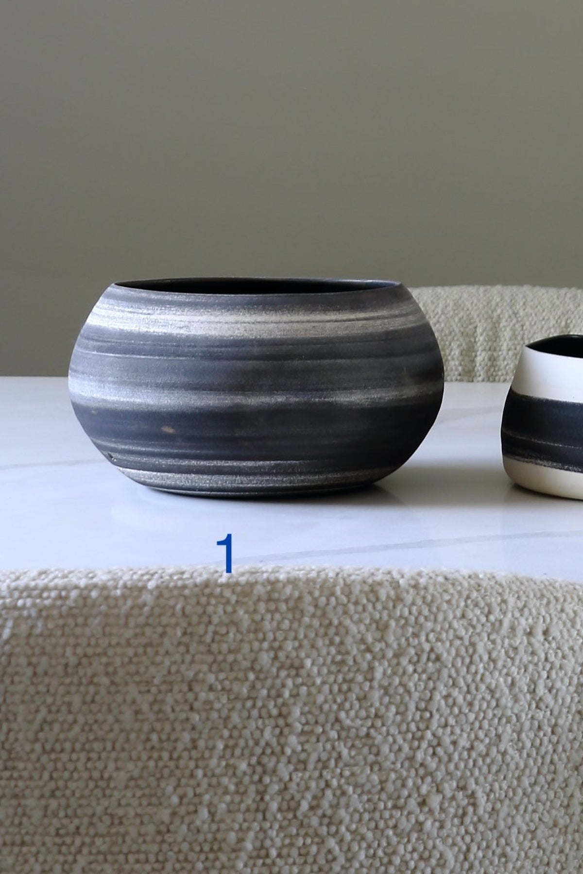 A round ceramic bowl with horizontal dark gray, black, and light gray glazed stripes, displayed on a white marble tabletop with a textured beige woven runner in the foreground.