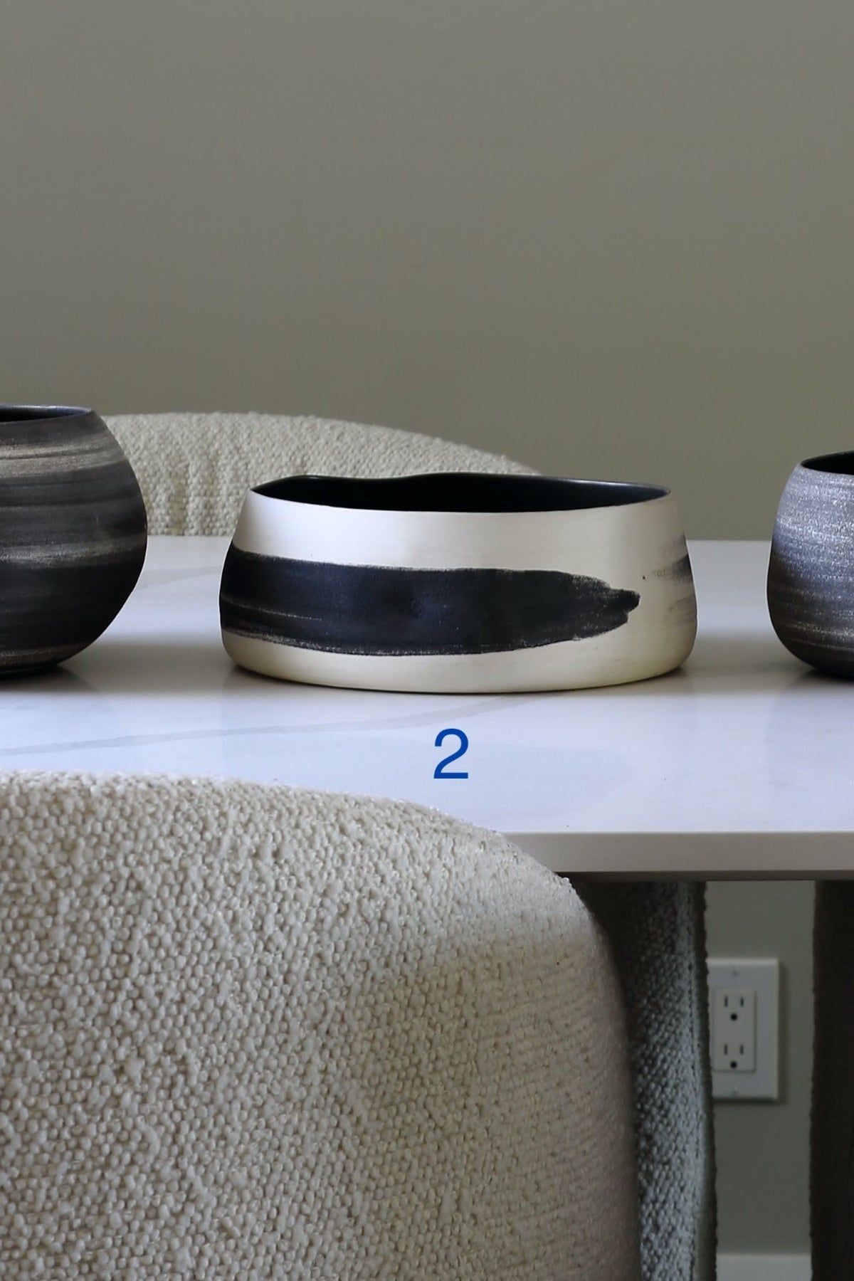 A wide, shallow ceramic bowl with a white exterior and a thick black brushstroke pattern on the front, displayed on a white table between textured beige chairs.