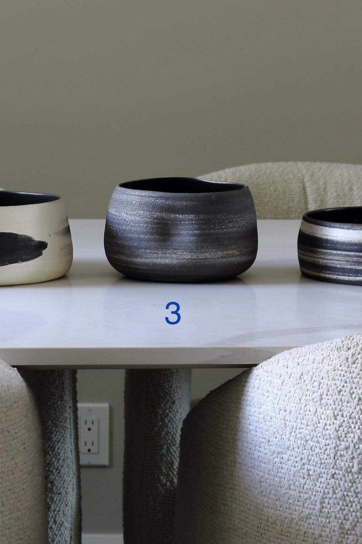 A dark gray and black textured ceramic planter pot displayed on a white table, with a blue number '3' printed below it. Other ceramic pots are partially visible on either side, and textured chairs are in the foreground.