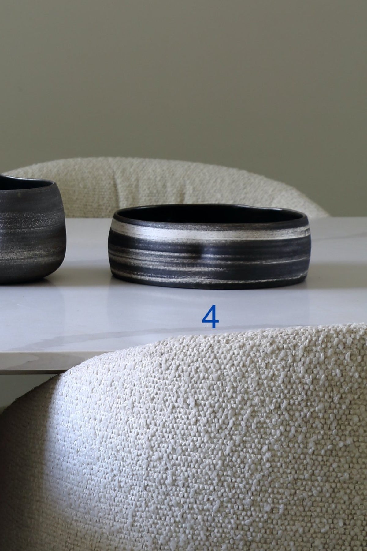 A wide, thick ceramic bowl with a black and white striped glaze sits on a white table next to another dark bowl, with a textured beige chair in the foreground and a neutral wall behind.