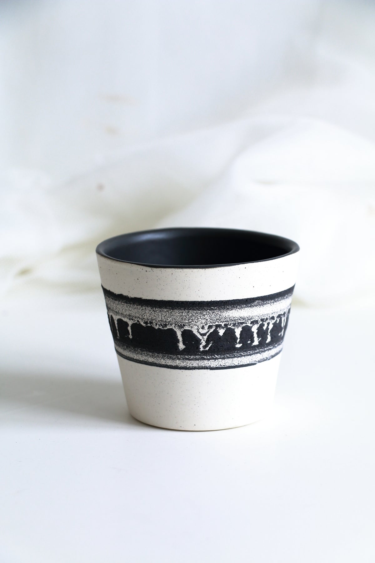 A small handmade ceramic cup with a black interior and a black dripping pattern band around the middle, set against a soft white fabric background.
