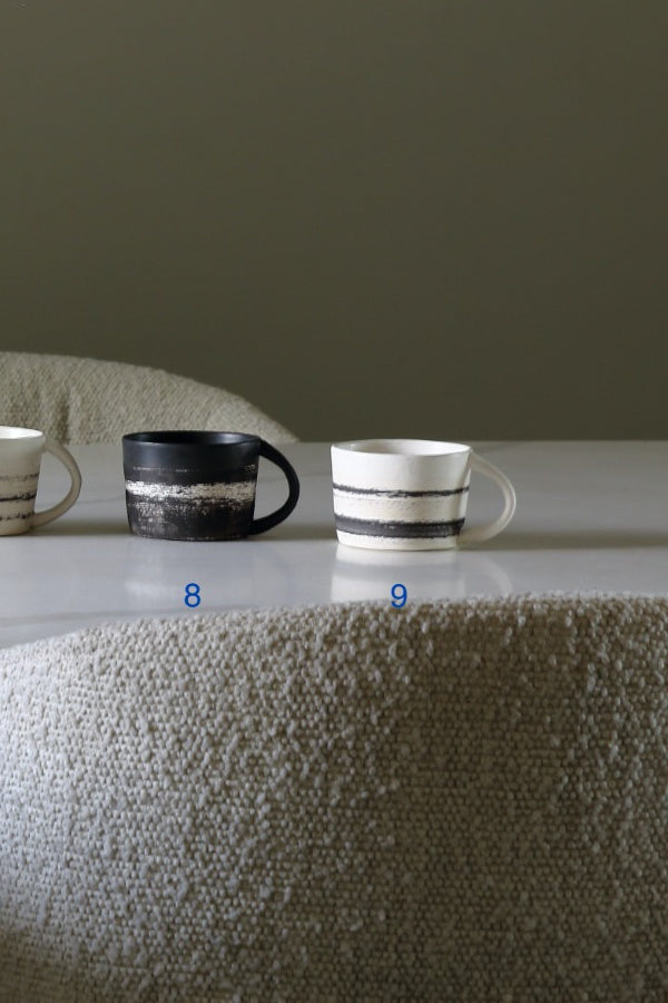 Two ceramic mugs displayed on a tabletop against a muted olive-gray background, with a textured beige chair in the foreground. One mug is black with a white band, and the other is white with black bands.