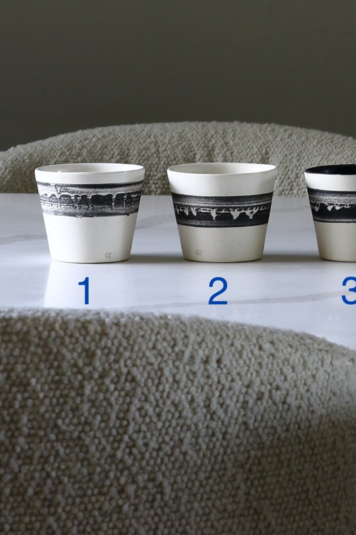 Three small white ceramic cups with black landscape-style bands near the rim, labeled 1, 2, and 3, displayed on a white surface in front of a textured beige chair.