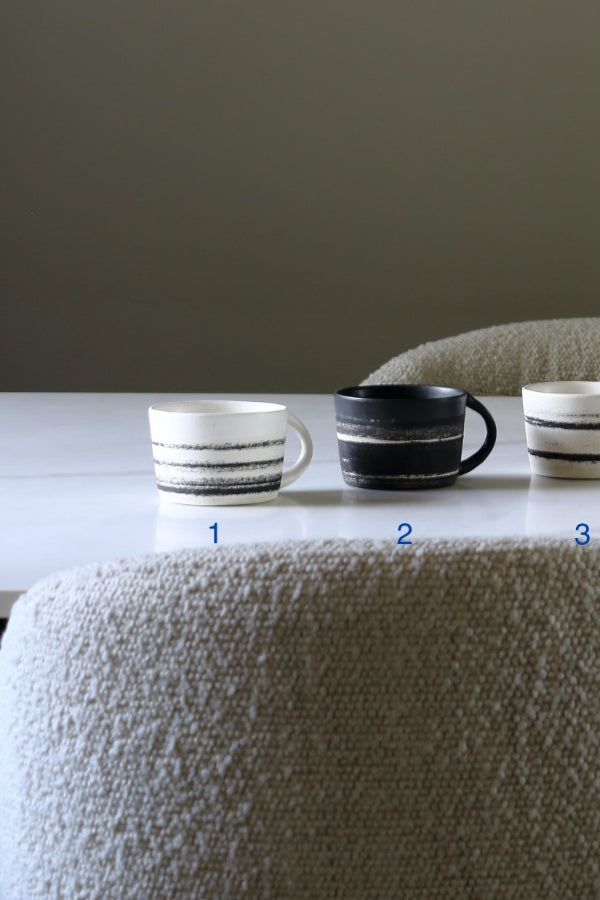 Three ceramic mugs with horizontal stripe patterns in black and white, arranged on a white table in front of a neutral gray background.