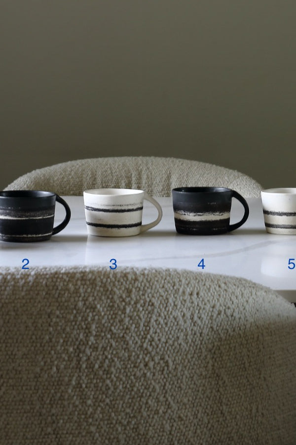 Five ceramic mugs with black and white horizontal stripe patterns are lined up on a white table in front of a textured beige chair. Blue numbers 2, 3, 4, and 5 are printed below each mug.