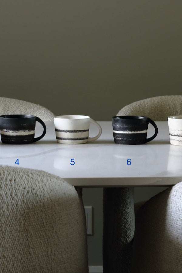 Four ceramic mugs in different color and pattern variations displayed on a white table in a dining setting with upholstered chairs. The mugs are numbered 4, 5, 6, and 7 in blue text.