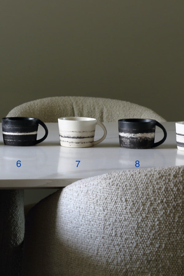 Four ceramic mugs displayed on a white table in front of upholstered chairs, with blue numbers 6, 7, 8 labeling them. The mugs are black and white with horizontal textured stripes.