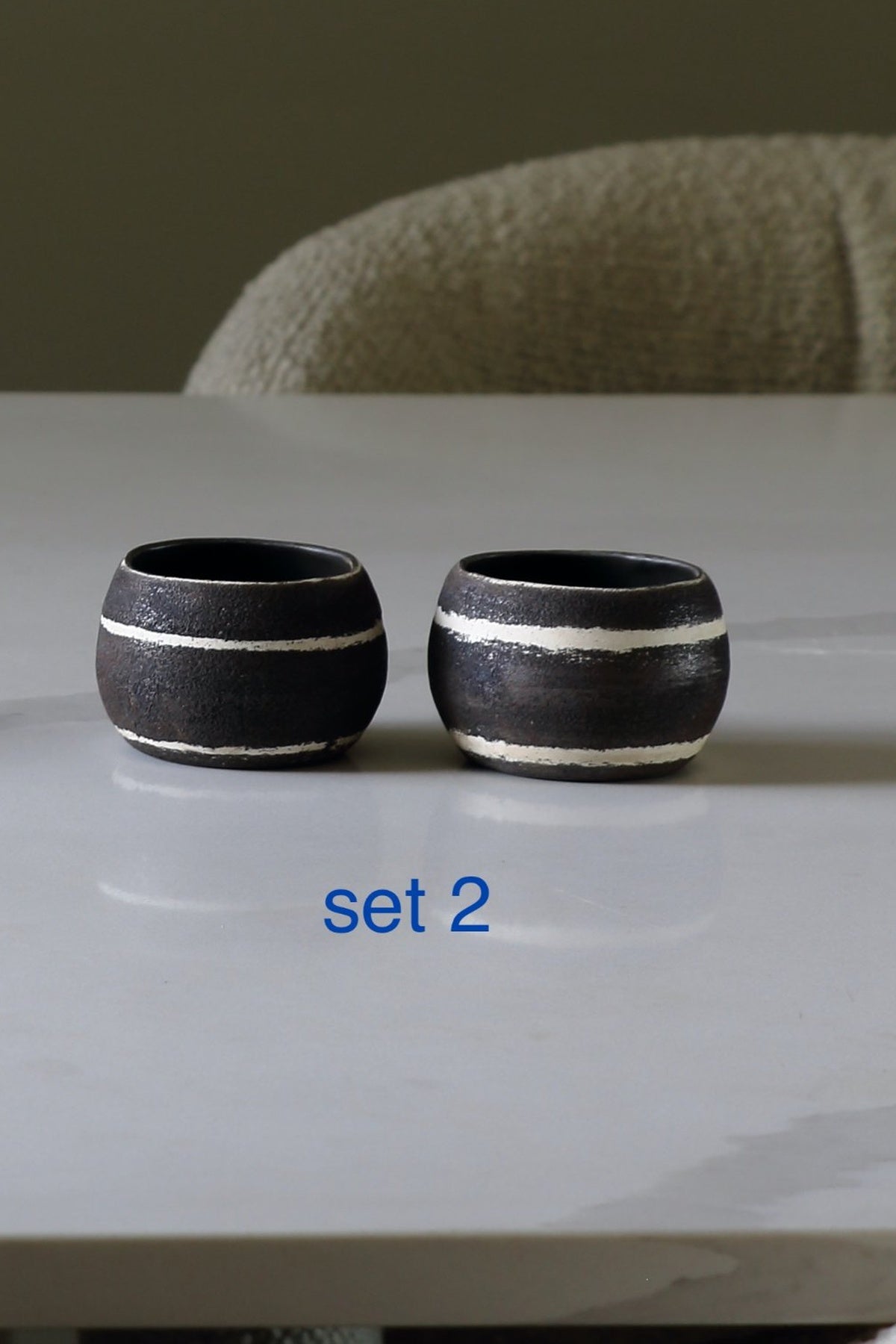 Two small round black ceramic cups with white horizontal stripes sit on a light gray tabletop, with a blurred chair in the background and the text 'set 2' below them.