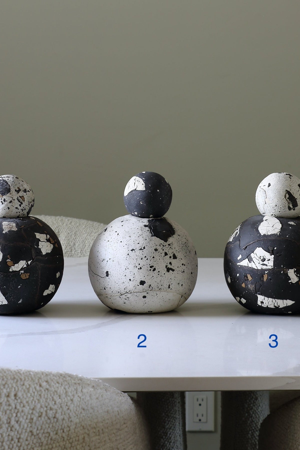 Three decorative ceramic globes with matching small rounded toppers sit on a white tabletop in an indoor setting. The left piece is black with white and gold speckling, the center piece is off-white with black speckling, and the right piece is black with white and gold speckling.