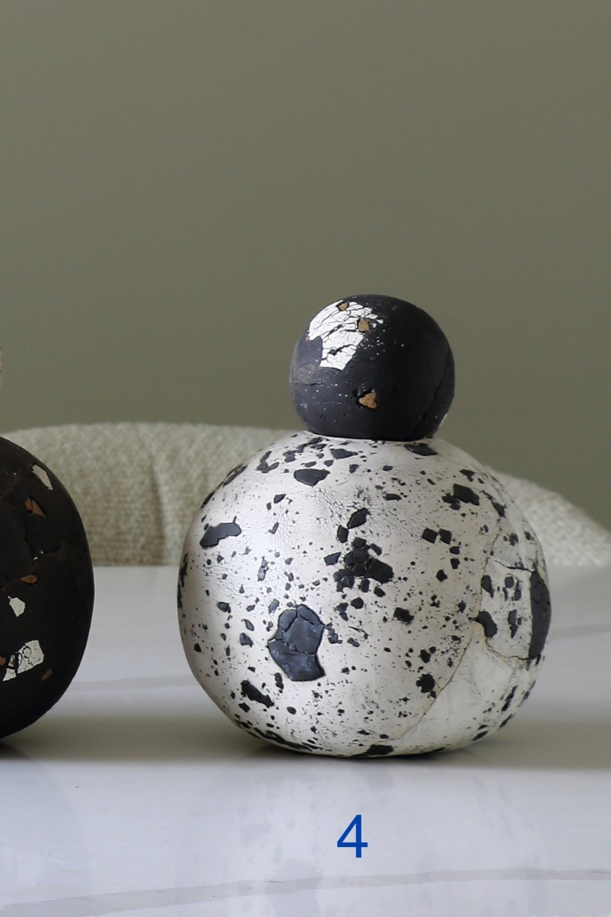 A decorative ceramic vase with a rounded white body covered in black speckles and a matching black top, shown on a light surface against a muted gray-green background. A partial second vase is visible at the left edge, and a blue number 4 appears below the main vase.