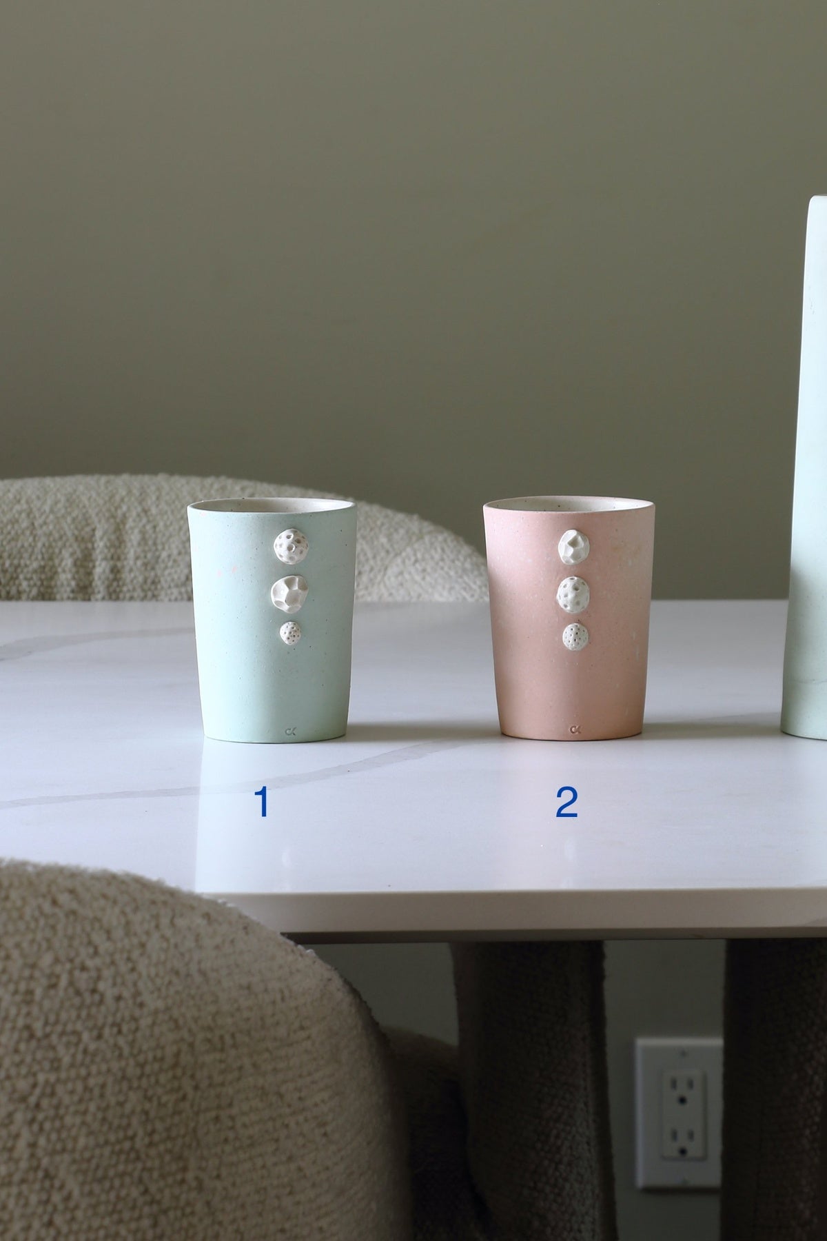 Two ceramic cups with textured white button details, labeled 1 and 2, sitting on a white table in a neutral indoor setting.